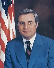 A man with dark hair, wearing a gray jacket, white shirt, and black striped tie