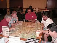 Five men sit at a table, poring over a map.  Dice and counters lie atop the map, some counters are stacked.  The map is subdivided into small hexagons of identical size.