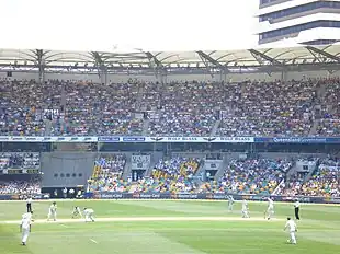 A bowler, midway through delivery. An umpire with black trousers and a light shirt watches from the left while the batsman not receiving the delivery is already walking down the pitch. In the background is a two-tier stand, full of spectators.