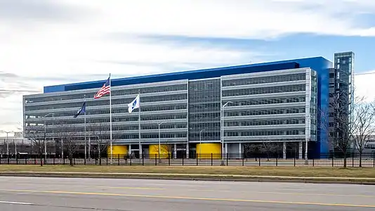 A wide rectilinear six-story blue-and-white building with the American, Canadian and Mexican flags flying in front, seen from a nearby roadway, under a cloudy sky