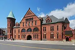 An ornate and complex dark red brick building, about three and a half stories tall, seen from across a wide street. It has a gabled front section, tall green-roofed conical tower on the left, and a lower engaged corner tower on the right.