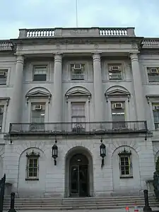 A section of a stone building. There is an arched entrance above steps at the lower level, and four columns leading to the top.