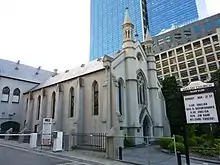 A grey stone church with skyscrapers in the background. The windows have pointed arches, there are two modest pinnacles on the main facade, which has an arched entrance and a traceried window above. A notice board states "Welsh Church", announces forthcoming services, and says "Welcome - Croeso!"