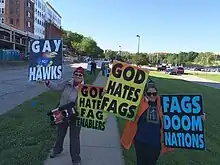 Two women standing on a university campus sidewalk, holding placards reading "Gay Hawks", "God Hates Fag Enablers", "God Hates Fags", and "Fags Doom Nations"