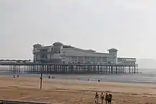 A walkway supported by metal legs arising from the sea, at the end of which is a large a white building with a wave-shaped roof and corner turrets.