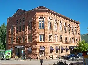 A three-story orange stone building on a slightly curved street corner, lit by sun from the right. The windows at street level have awnings; those at the top are in rounded arches. The face of the building on the left has a small pointed top at the roof. At street level an awning on that face has "Wheeler Opera House" on it; the word "Bank" is carved into the stone on the corner face above an awning with "Bentley's" written on it.