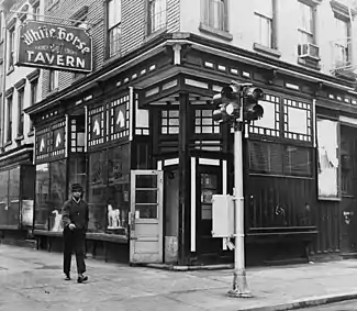  On the corner of a block is a building with large glass fronts on both sides; a sign displaying the tavern's name shines brightly above in red neon.