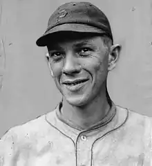 A man wearing a lightly colored baseball uniform holds his hands above his head, winding up to throw.