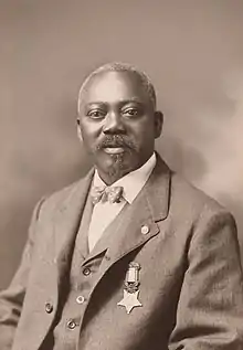 A black man with beard seated, wearing a tweed suit with the Medal of Honor pinned to it.