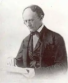 An older man with a receding hairline and reading glasses looks down at the book he is reading. He wears a dark suit with an old fashioned bow-tie