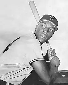 A man, wearing a baseball cap with an interlocking "SF" logo, clutches a baseball bat and prepares to swing.