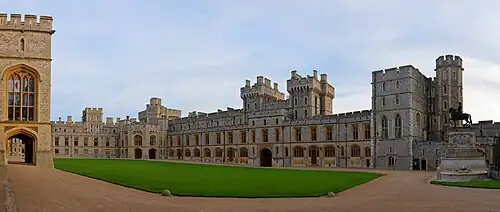 A photograph of a grey Gothic quadrangle with a green grass square in the middle. On the left, a block of the building makes up the near ground. A gatehouse is in the middle of the right hand part of the quadrangle.