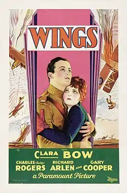 The theatrical poster of Wings. It focuses on two people. Buddy Rogers is wearing an aviator suit and Clara Bow is wearing a jacket and gloves.