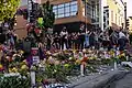 People gather around piled flowers, candles, and signs memorializing Knajdek