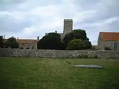 Square stone tower and red roofed buildings behind a stone wall and partially obscured by trees.