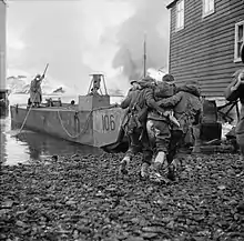 A black and white photograph of a wounded soldier being helped to a landing craft by two other soldiers