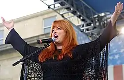 A woman with long red hair wearing black, singing into a microphone while raising both her arms