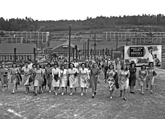 Workers, mostly women, pour out of a cluster of buildings. A billboard exhorts them to "Make C.E.W. COUNT continue to protect project information!"