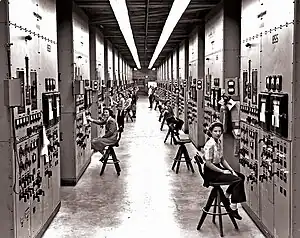 A long corridor with many consoles with dials and switches, attended by women seated on high stools