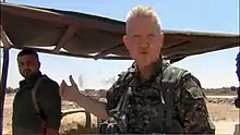 Michael Enright wearing army uniform, squinting and appearing to speak directly to camera, standing under makeshift shelter with smoke in the background and other armed men next to him