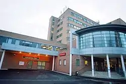 Multi-storey building with long lines of windows on each floor. On the roof are various aerials. In the foreground are cars on a roundabout and trees.