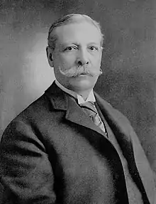 A light-haired gentlemen with a handlebar moustache in a dark coat and wing collar