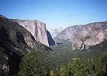 A glacial-carved valley filled with evergreen trees has a hanging valley and waterfall on the right and high cliff face on the left.