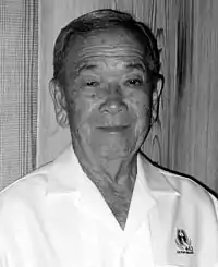 Head and shoulders of an elderly man wearing a white button shirt with an emblem on the left breast bearing the text "442" and "GO FOR BROKE".