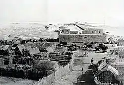 Photo of a settlement with thatched-roof buildings and people walking, seen from above