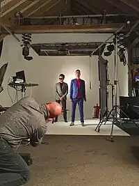 Two members of Zoot Woman stand amidst white panels, surrounded by camera equipment. A third man, on his knees, tilts his head looking toward the Zoot Woman members